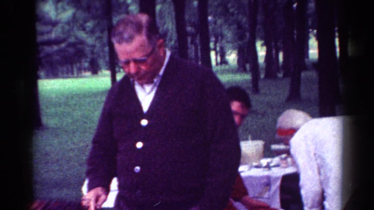 An older man in a dark cardigan stands in the foreground while adults work around a picnic table in a wooded Toledo park setting.