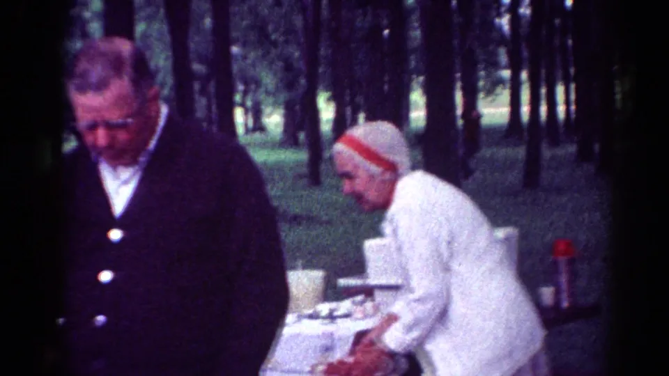 An older woman in a light sweater and knit hat bends over a picnic table while an older man stands nearby in 1964 Toledo park footage.