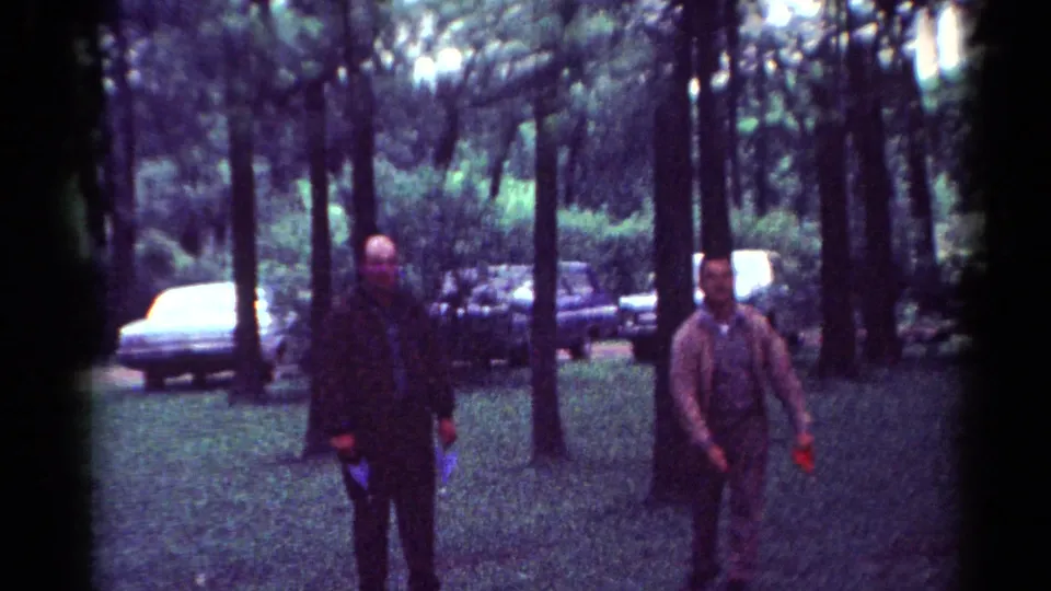 Two men walk through trees in a Toledo park, with mid-century cars parked behind them in this 1964 home-movie frame.