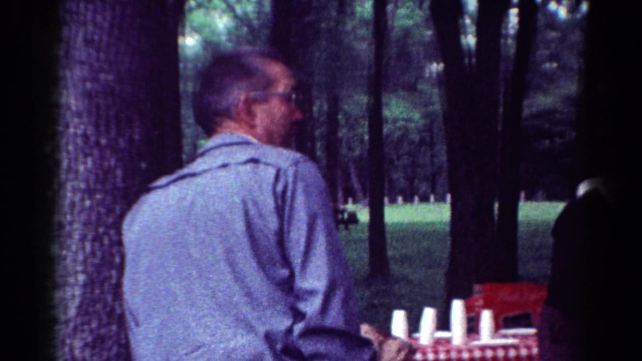 Man in a light blue shirt stands beside a checkered picnic table in a wooded park.
