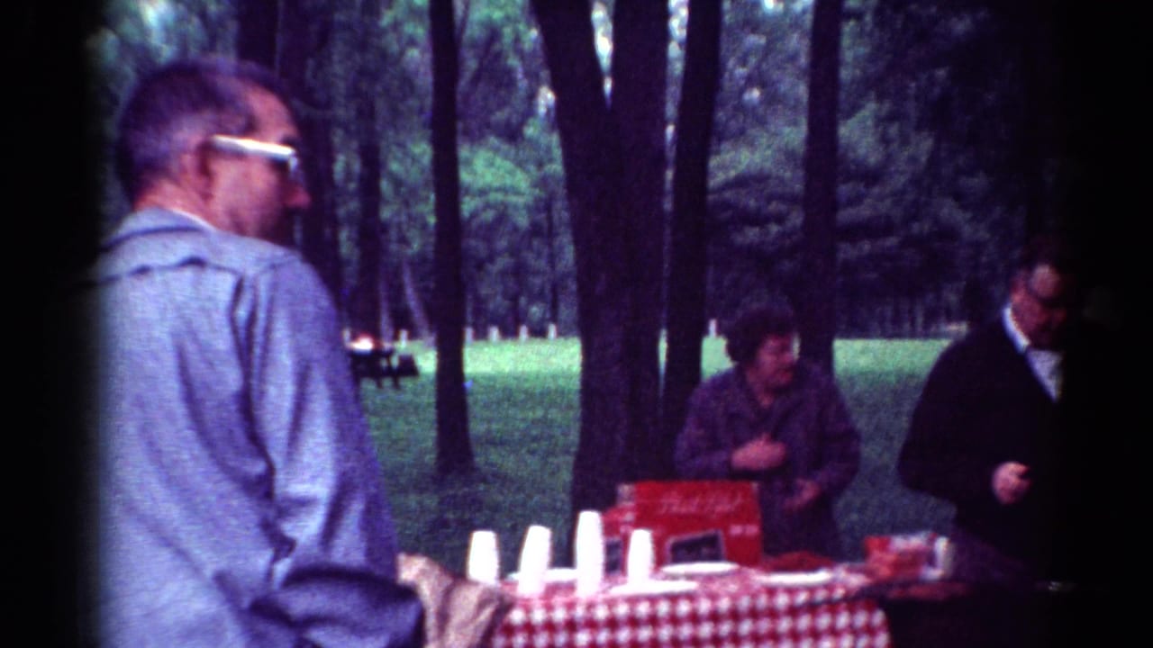 Adults gather near a checkered picnic table with cups and red cartons in the trees.