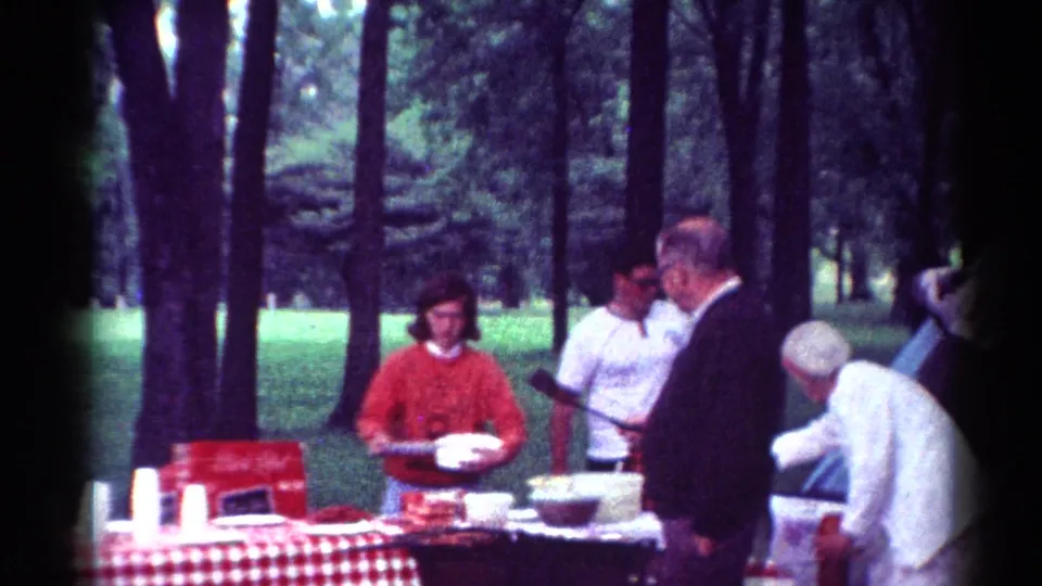 Adults gather beside the picnic table in a wooded Toledo park, with one person holding a plate and cups and bowls set out nearby.