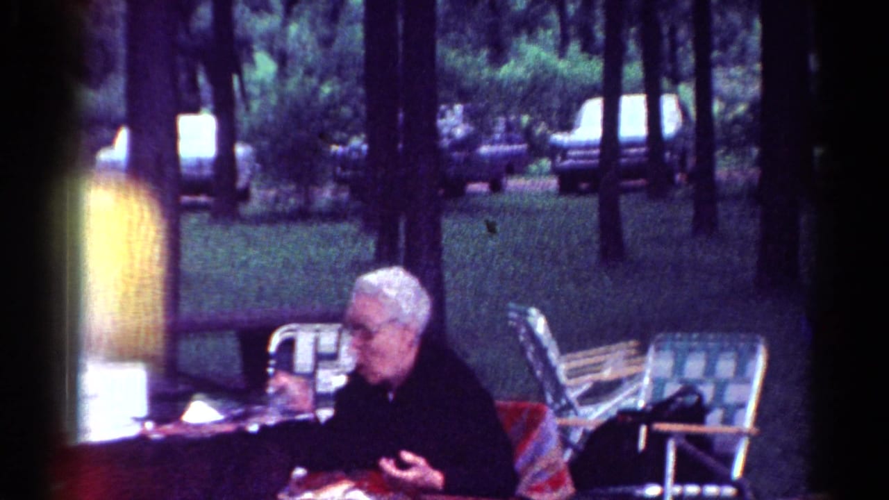 Elderly woman with white hair and glasses sits at a picnic table in a wooded park.