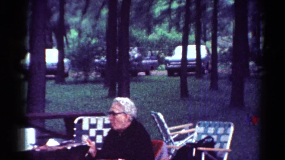 The woman remains seated at the picnic table in the wooded Toledo park setting, with patterned lawn chairs and cars in the background.