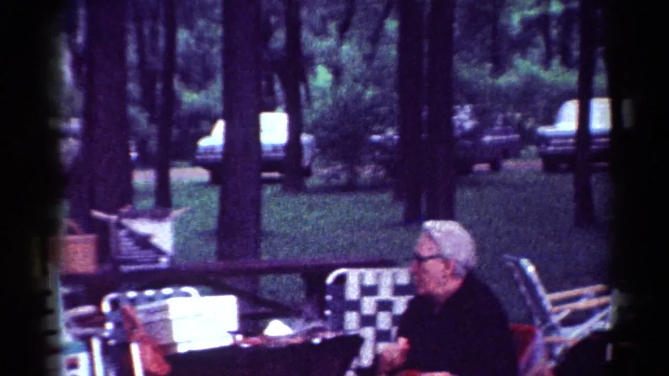 The 8mm frame shows the elderly woman at the outdoor table, surrounded by folding chairs, trees, lawn, and distant cars.