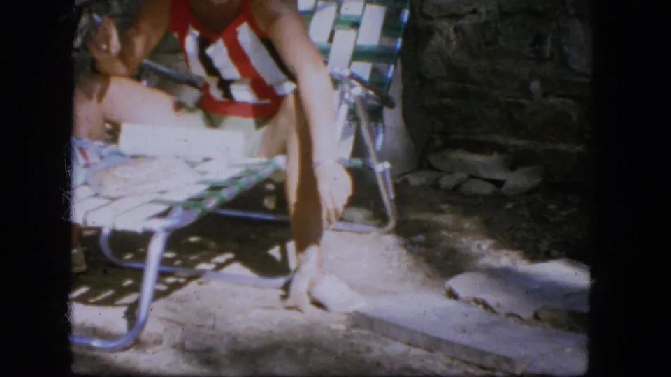 1964 Troy frame showing a person in a striped swimsuit seated beside a stone wall while a small animal stays near the chair.