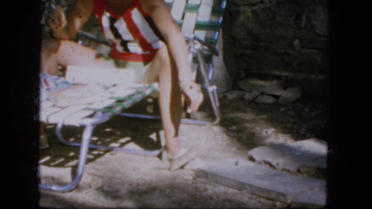 1964 Troy, New York home-movie frame showing a rodent beside a seated woman's bare feet on a sunlit stone patio.