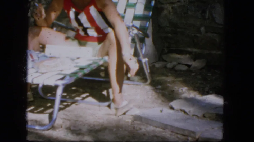 1964 Troy, New York home-movie frame showing a rodent beside a seated woman's bare feet on a sunlit stone patio.