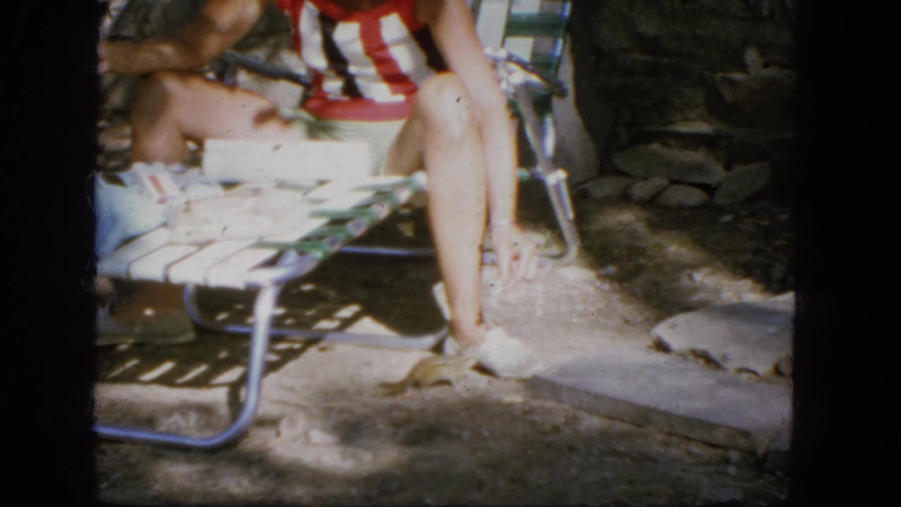 1964 Troy, New York home-movie frame with a seated person in a red-and-white striped shirt on a metal lawn chair near stonework.