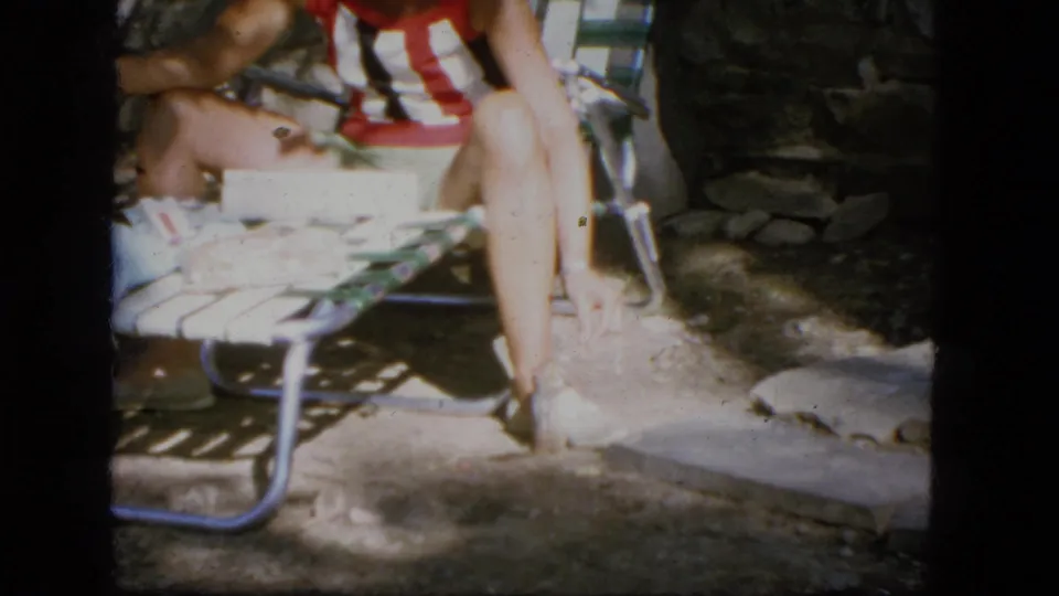 1964 Troy, New York frame showing a seated person on a low lawn chair, with striped shirt, legs, stonework, and patio ground visible.