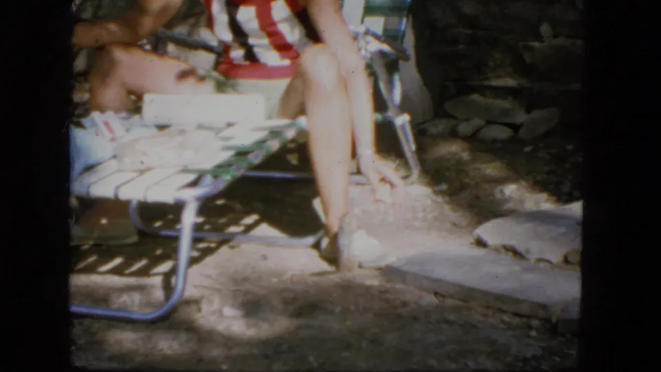 1964 Troy, New York 8mm frame of a seated person on a webbed lawn chair near a stone wall and cabin-yard patio.