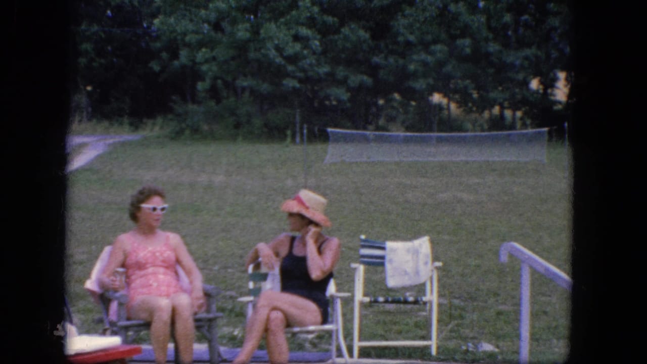 Two women in vintage swimwear sit on a concrete poolside patio in Troy, New York, with lawn chairs, a towel, grass, trees, and a tennis net behind them.