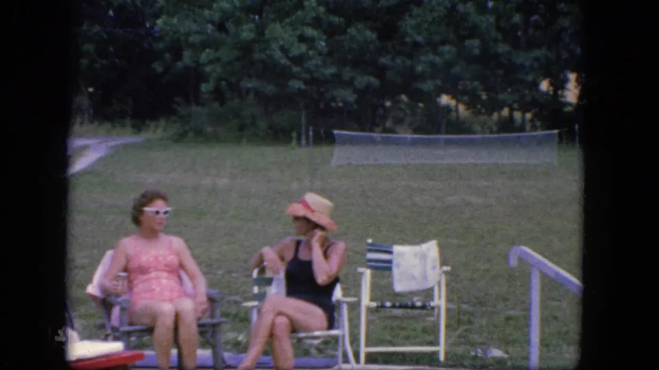 Two women in vintage swimwear sit on a concrete poolside patio in Troy, New York, with lawn chairs, a towel, grass, trees, and a tennis net behind them.