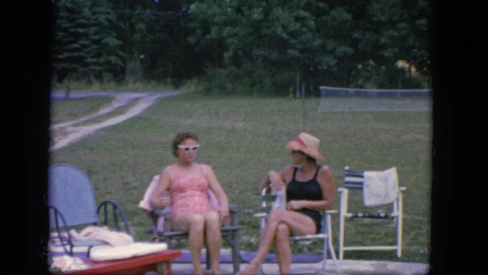 A 1960s home-movie frame shows two women seated beside a pool area in Troy, with a lounge chair at left and lawn, trees, and a tennis net behind them.