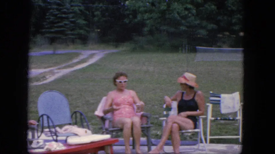 In a Troy, New York 8mm frame, two women in swimwear sit in folding chairs by a poolside patio, with one gesturing during conversation.