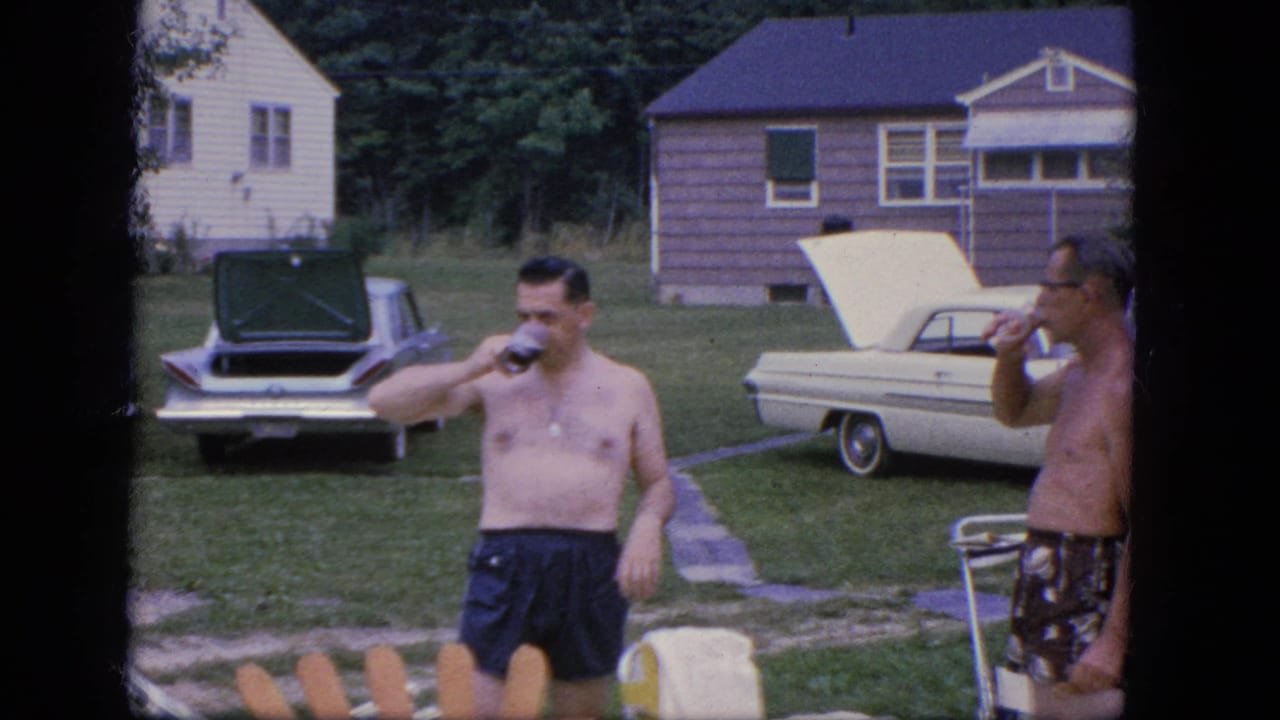 Two shirtless men in swim trunks stand in a yard with bottles and parked cars behind them.