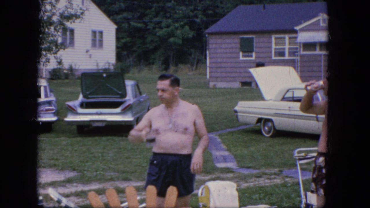 A shirtless man holds a bottle in a yard while another man is partly visible at the edge.