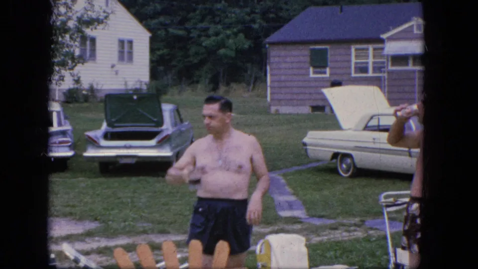 A shirtless adult man stands in a Troy backyard with a bottle as parked cars and small houses appear in the 1964 home-movie frame.