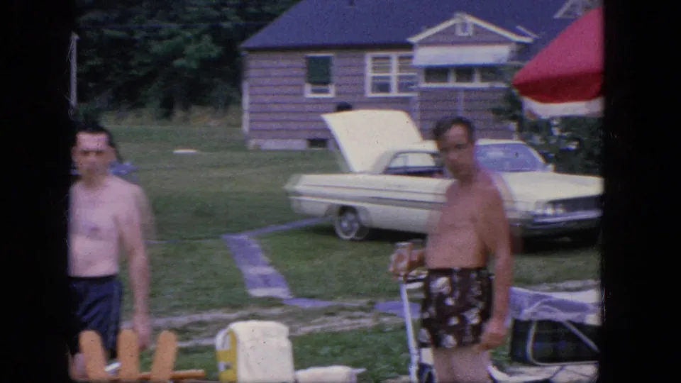 Two adult men in swim trunks move through a 1964 Troy backyard with lawn chairs, a red umbrella, and a white sedan nearby.