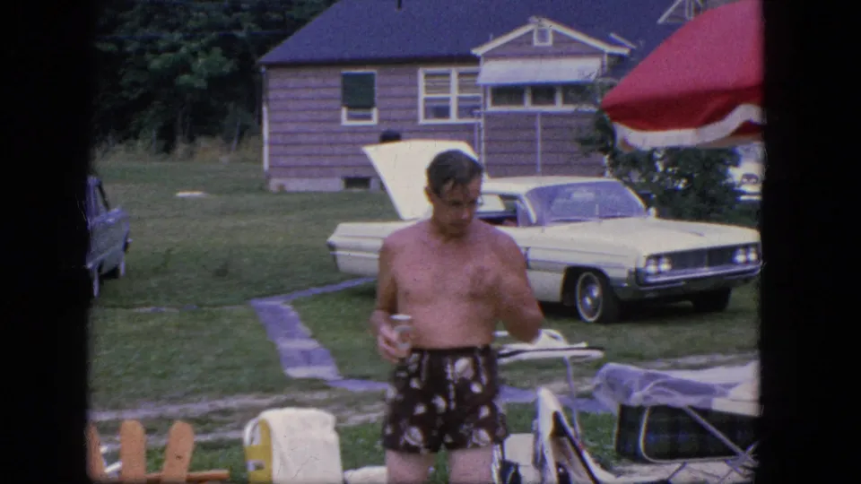 A shirtless adult man in patterned swim trunks holds a cup in a Troy backyard, with a white sedan and lawn chair behind him.