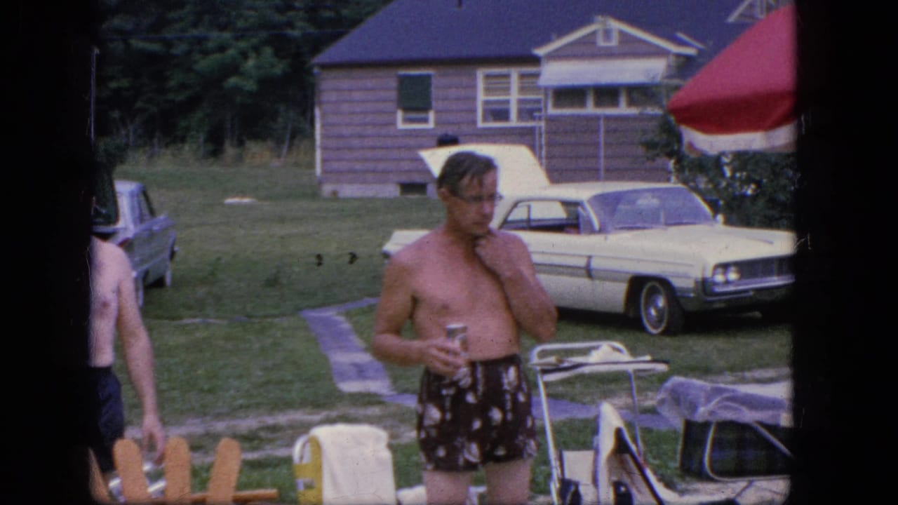 Shirtless man in patterned swim trunks holds a drink in a grassy yard near a white sedan.