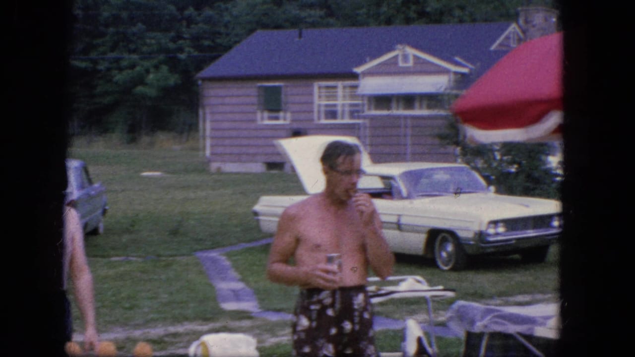 Man in swim trunks holds a drink and raises a snack near lawn furniture and a parked car.