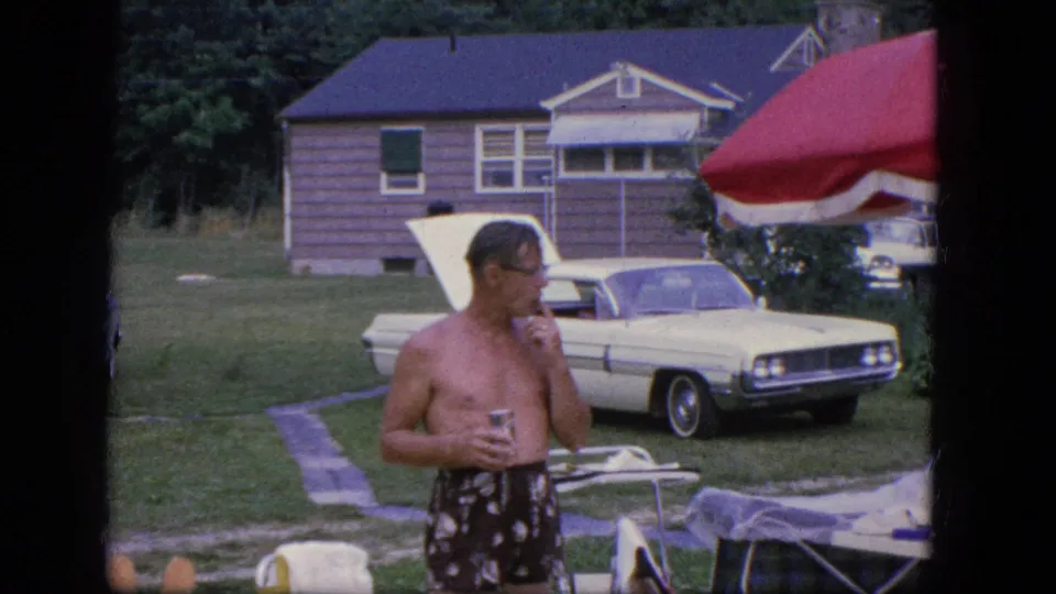 A shirtless man in patterned swim trunks looks downward in the yard, with a white sedan and wooden house in the background.