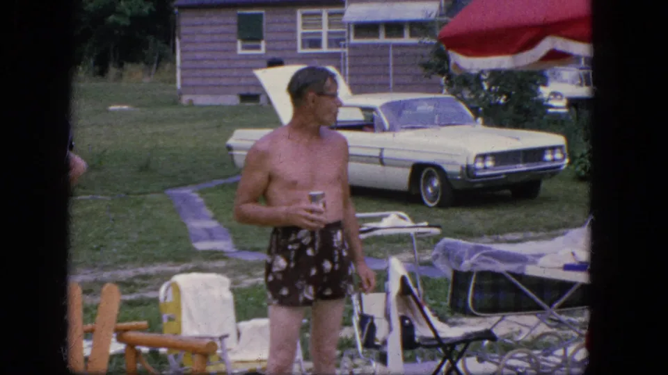 The man stands in a grassy Troy yard holding a drink, with lawn furniture, a red-and-white umbrella, and a white sedan nearby.