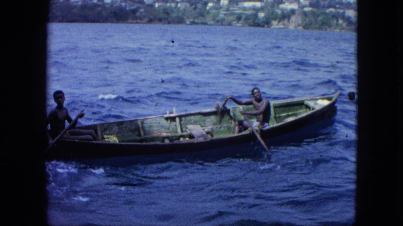 Two men row a small wooden boat across choppy blue water near a distant shoreline.