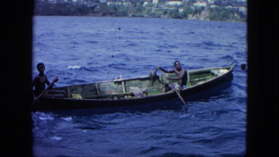 1964 U.S. Virgin Islands 8mm frame showing two men in a wooden rowboat on blue water with green shoreline in the distance.
