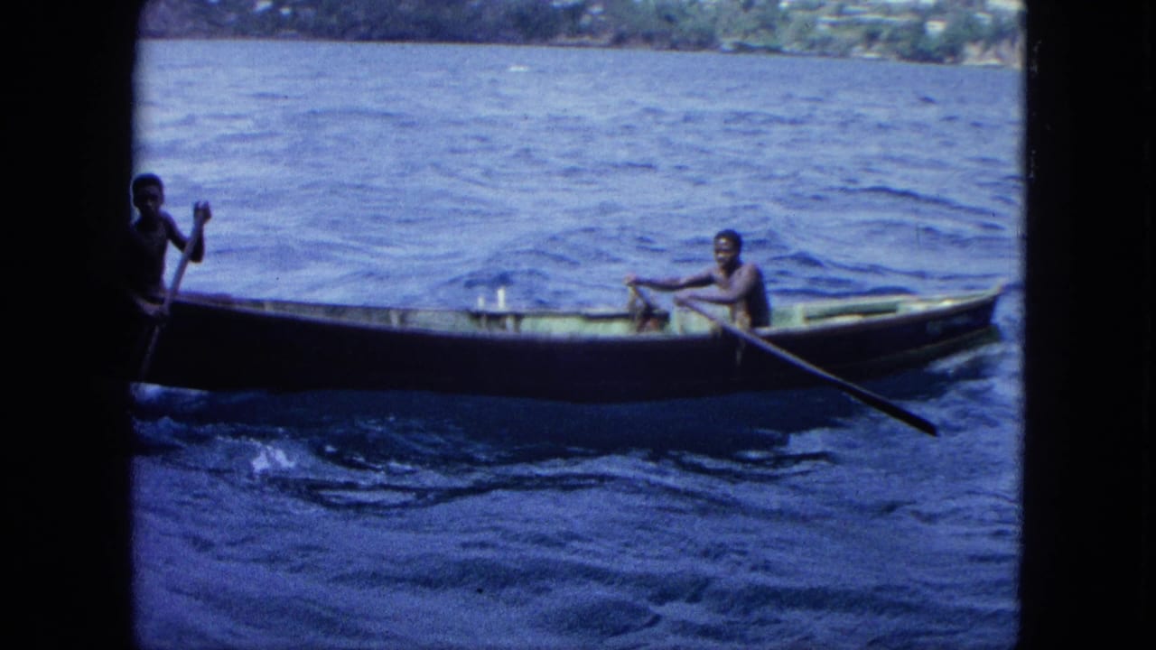 A wooden rowboat passes across blue water with two men aboard and shoreline behind it.