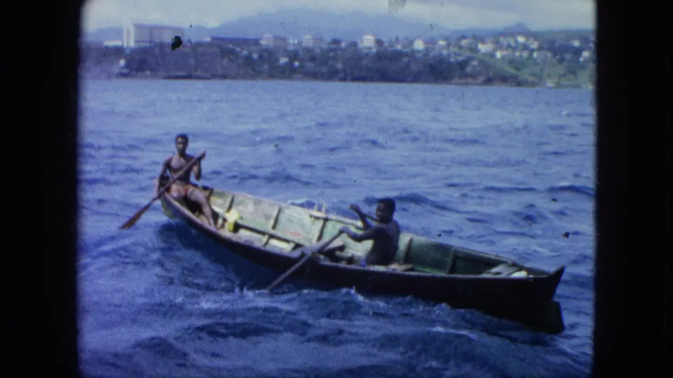 1964 U.S. Virgin Islands 8mm frame showing a small wooden boat angled through choppy water, with two men using oars and shore in the background.