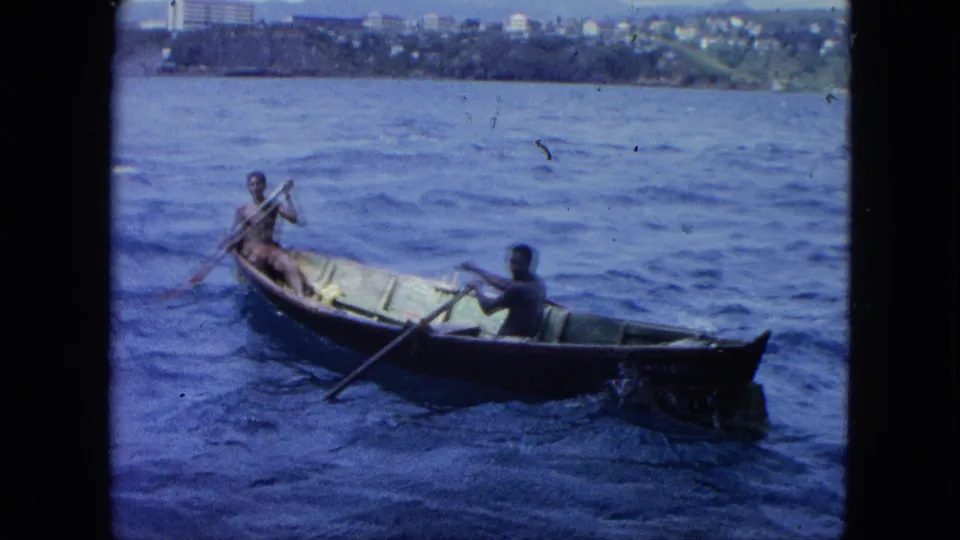 1964 U.S. Virgin Islands frame of two men rowing a wooden boat across open water, with a low shoreline and scattered buildings in the distance.