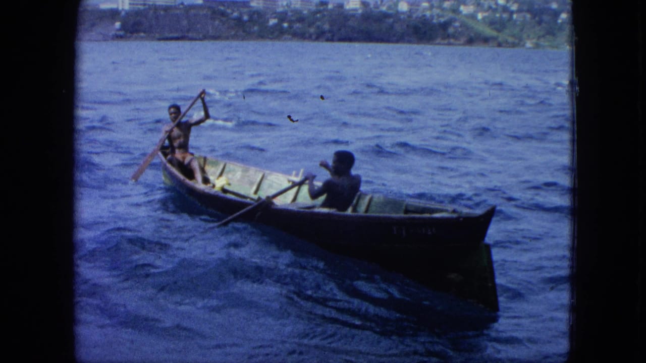Two men row a small wooden boat across choppy blue water near a distant shoreline.