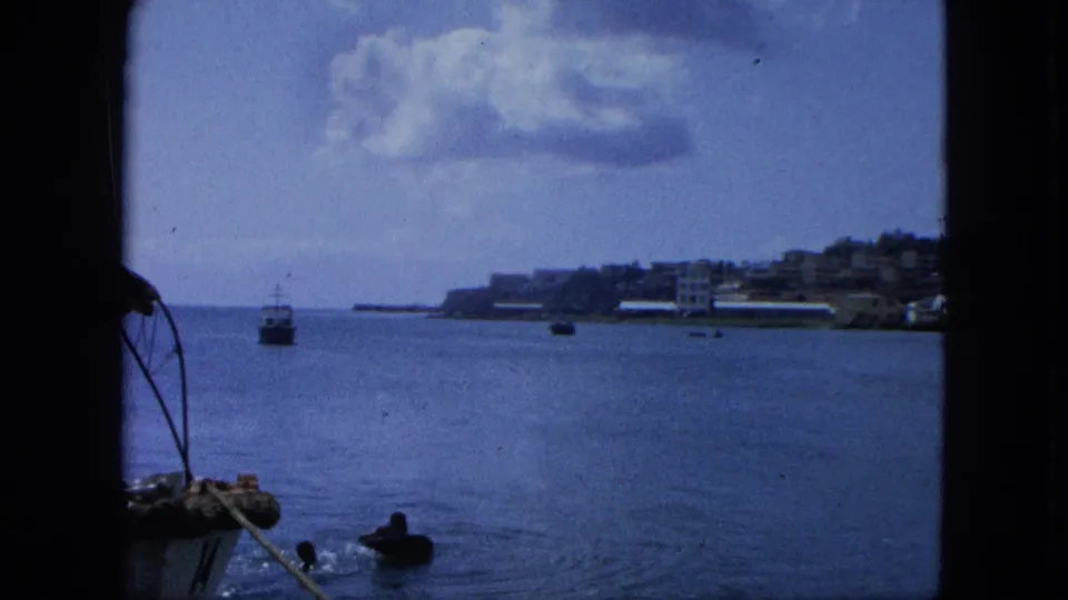 1964 United States Virgin Islands 8mm frame showing a wider harbor view with boats, shoreline buildings, and clouded sky.