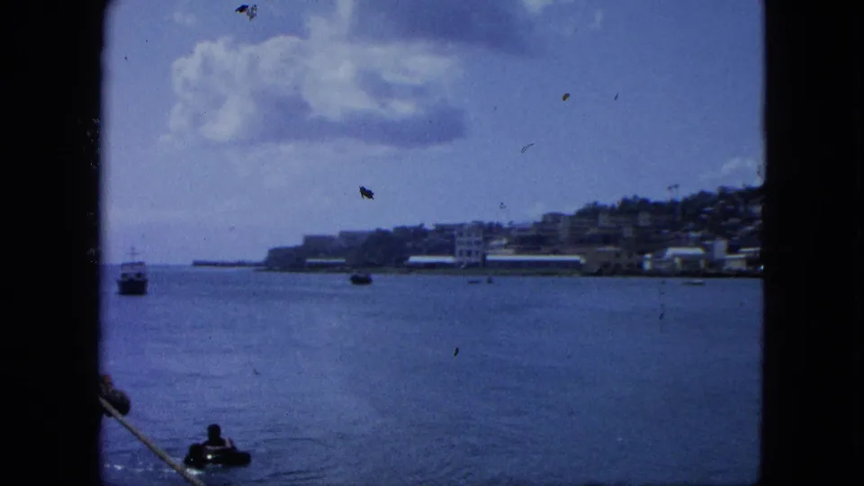 1964 U.S. Virgin Islands 8mm frame showing blue water, distant shoreline buildings, hillside, clouds, and a dark vessel-frame edge.