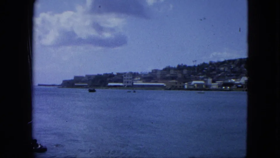 1964 U.S. Virgin Islands coastal view with calm blue water, small boats, distant buildings, hillside, and soft clouds.