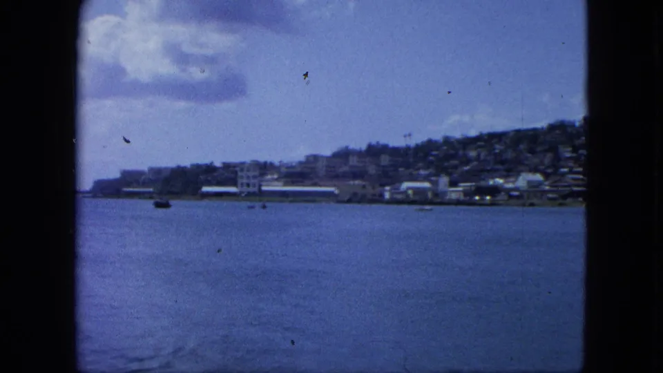 1964 8mm travel frame of U.S. Virgin Islands shoreline, boats on blue water, hillside buildings, clouds, and gate edges.