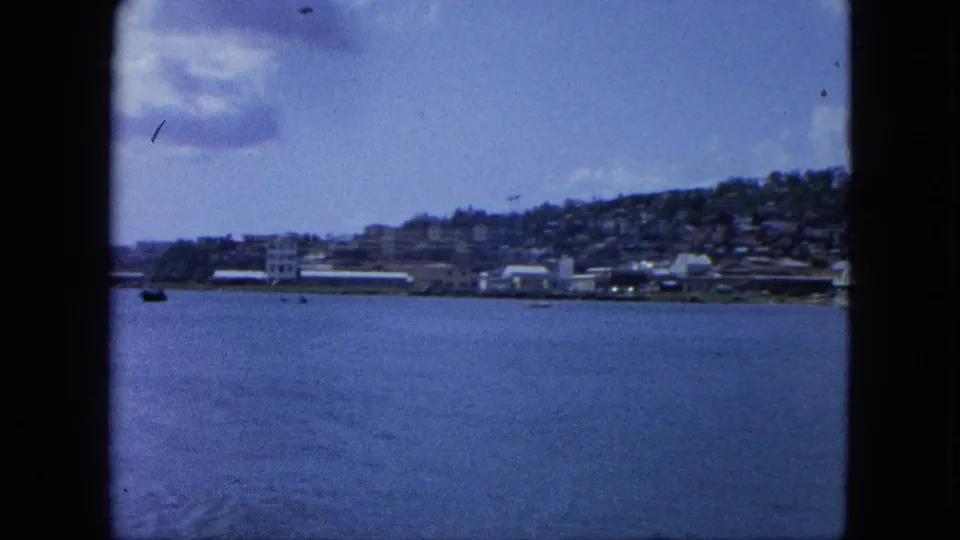 1964 U.S. Virgin Islands home-movie frame with calm water, distant shoreline, hillside structures, small boats, and blue sky.
