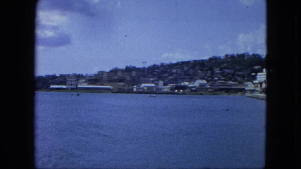 1964 U.S. Virgin Islands waterfront view with calm water, low shoreline buildings, and a hillside town in soft 8mm color.