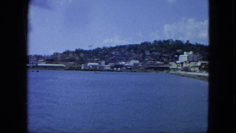 1964 U.S. Virgin Islands shoreline with blue harbor water, low coastal buildings, hillside housing, and visible 8mm frame edges.
