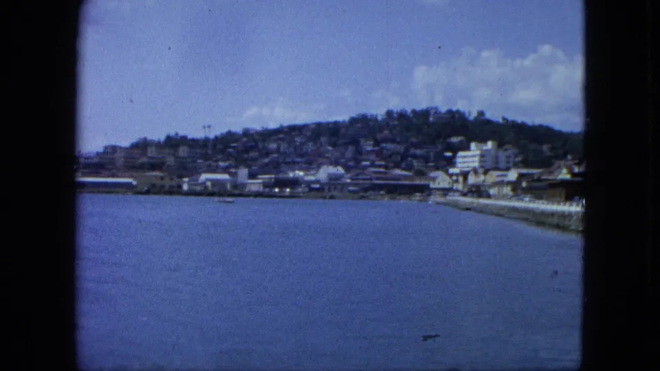 1964 U.S. Virgin Islands coastal view with a seawall at right, waterfront buildings, calm water, and hillside homes beyond.