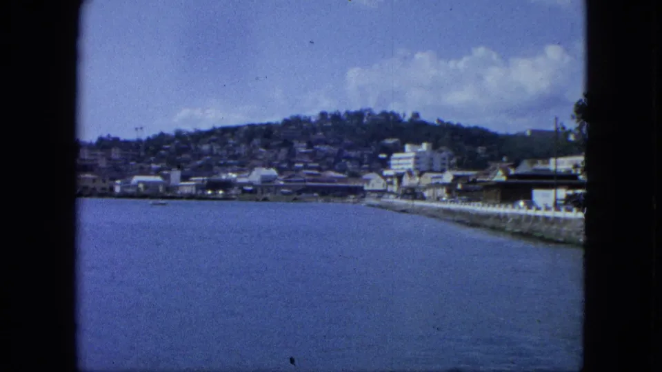 1964 U.S. Virgin Islands waterfront with blue water in the foreground, a seawall, low buildings, and a hillside town under clouds.