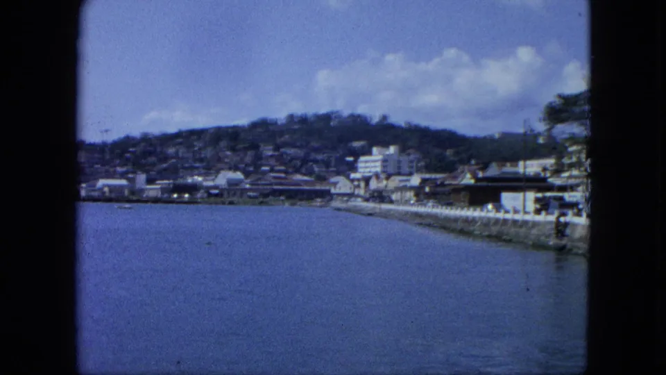 1964 8mm view toward a USA Virgin Islands waterfront, with calm water, a seawall street, hillside housing, and scattered clouds.