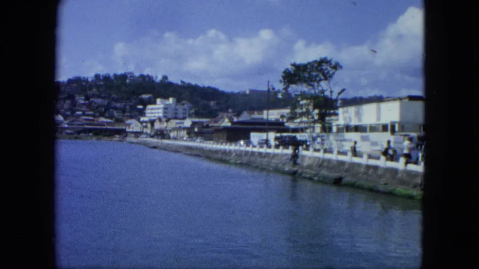 1964 8mm frame of a USA Virgin Islands waterfront street, showing pedestrians and cars near a seawall below hillside homes.