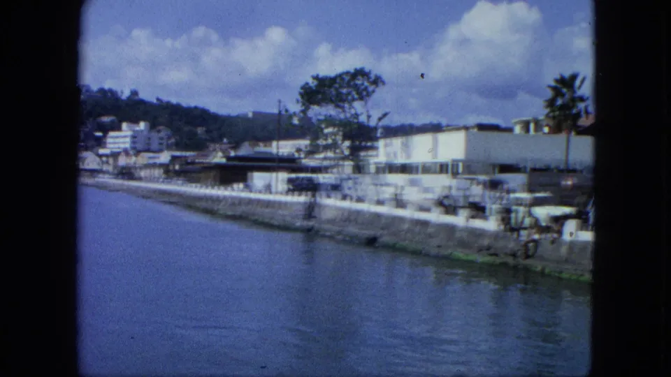 1964 USA Virgin Islands waterfront seen from the water, with buildings, seawall, trees, cars, and hillside development beyond.
