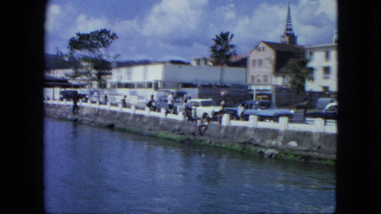 Waterfront road with pedestrians, classic cars, palm trees, buildings, and a church steeple.