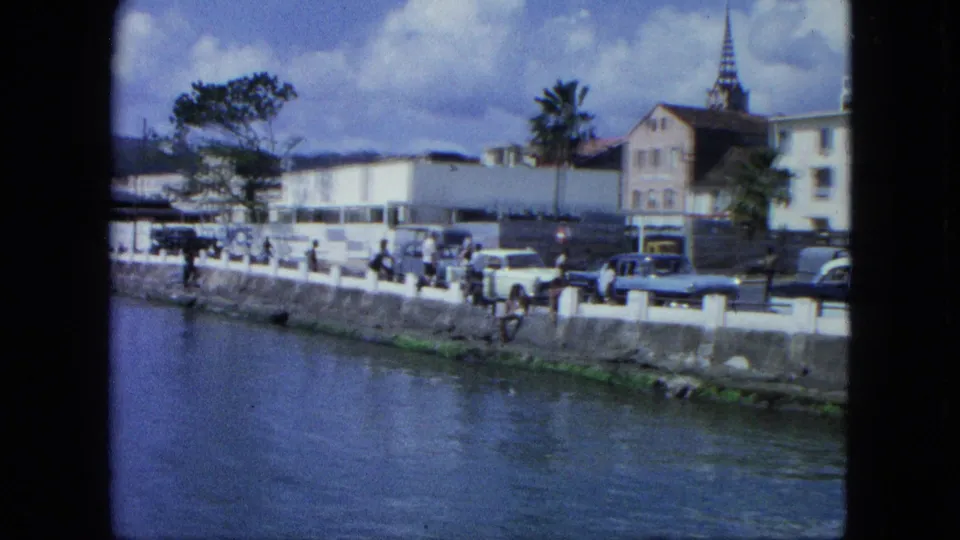 1964 USA Virgin Islands harbor street with people along the seawall, mid-century cars, palm trees, buildings, and a church steeple.