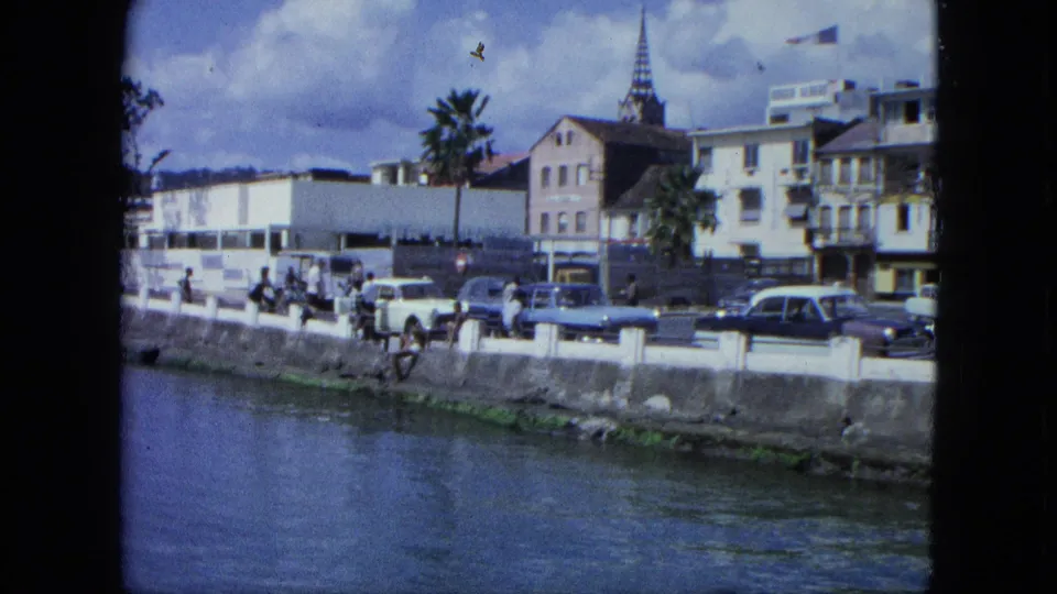 1964 USA Virgin Islands waterfront scene showing pedestrians near a seawall, period cars, palm trees, hotel signage, and a church steeple.