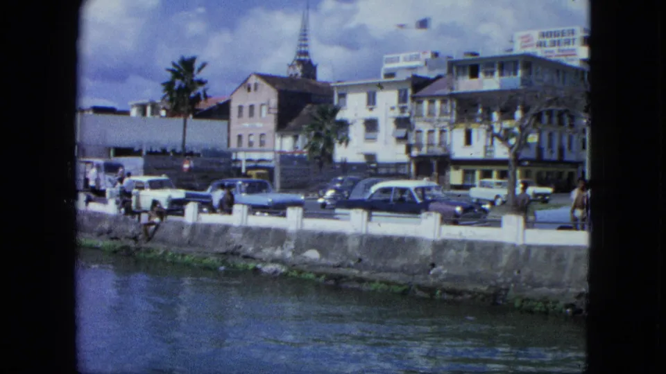 1964 USA Virgin Islands harborfront view with a blue classic car, pedestrians, palm trees, hotel signage, waterfront buildings, and a church steeple.